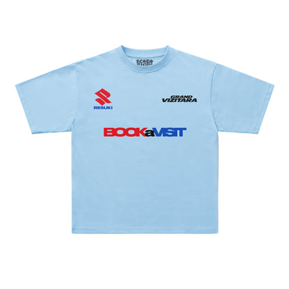 A light blue short-sleeve t-shirt with sponsor logos printed on the chest, including Suzuki, Bock, and Grand Vizita, displayed flat against a light gray background.