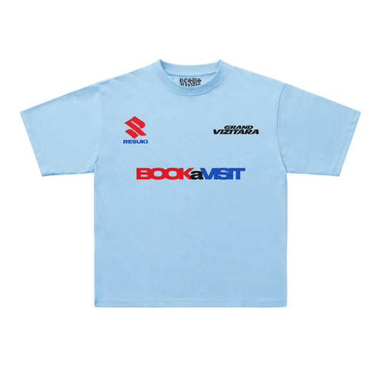 A light blue short-sleeve t-shirt with sponsor logos printed on the chest, including Suzuki, Bock, and Grand Vizita, displayed flat against a light gray background.